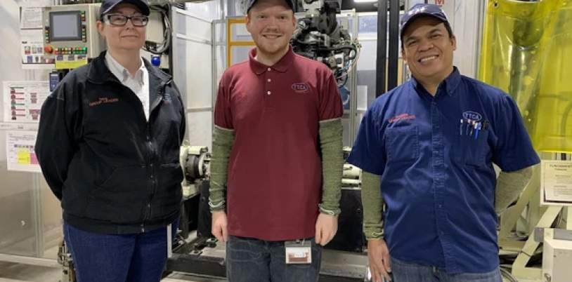 Alex stands in between two other adults in front of some factory equipment