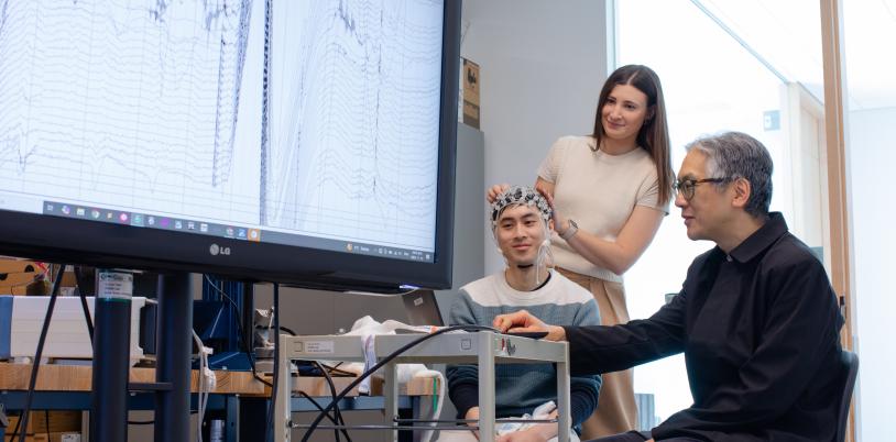 An adult sits and wears a brain computer interface headset while two other adults look at brain wave imaging.