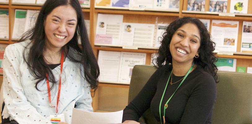 Two women with dark hair hold papers with shelves behind them