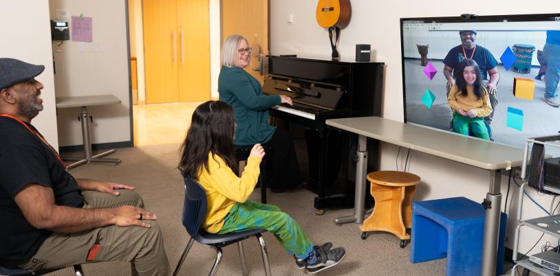 Child in yellow shirt sits in front of screen with icons with a woman on piano beside her and a man sitting behind her