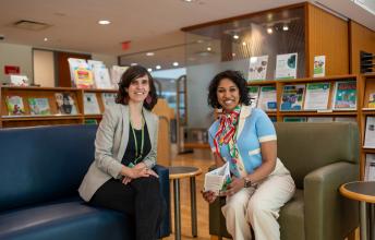 Two adults sit on arm chairs in the library, surrounded by shelves of books and pamphlets.