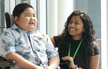 Boy with dark hair smiles in wheelchair beside woman with long dark hair who is also smiling