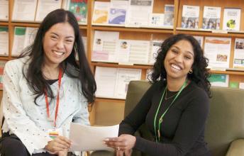 Two women with dark hair hold papers with shelves behind them