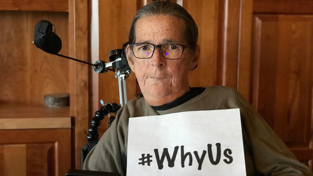 Woman in wheelchair holding sign that says #WhyUs