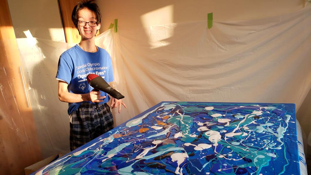 Teenage boy beside canvas with blue shades of paint poured over it holding blow dryer