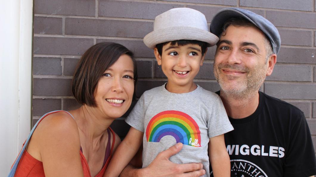 A woman with short brown hair is smiling and hugging her young son. The young boy is smiling and wearing a grey t-shirt with a rainbow on it. His father is kneeling beside him and holding him. He is wearing a black t-shirt and a baseball cap.