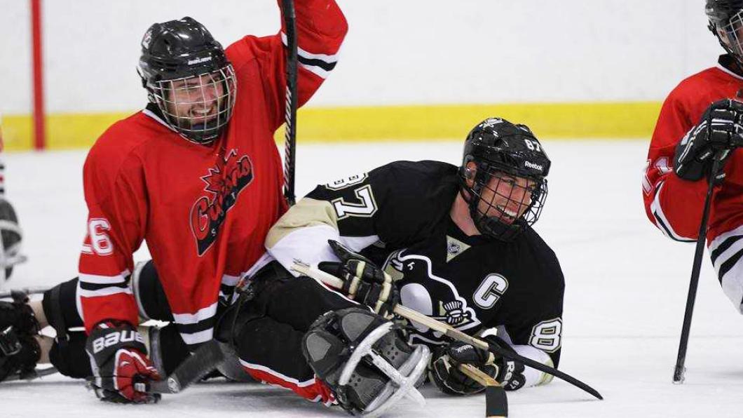 Photo of two sledge hockey players on the ice one with red jersey and one with black