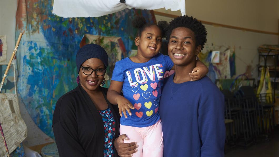 An adult with deep skin tone wearing a blue head-wrap, purple glasses and a black shawl. They are standing next to a young child with deep skin tone, who is wearing a blue t-shirt and pink pants. The child's arm is around a teenager, who has deep skin tone and is wearing a blue sweater.