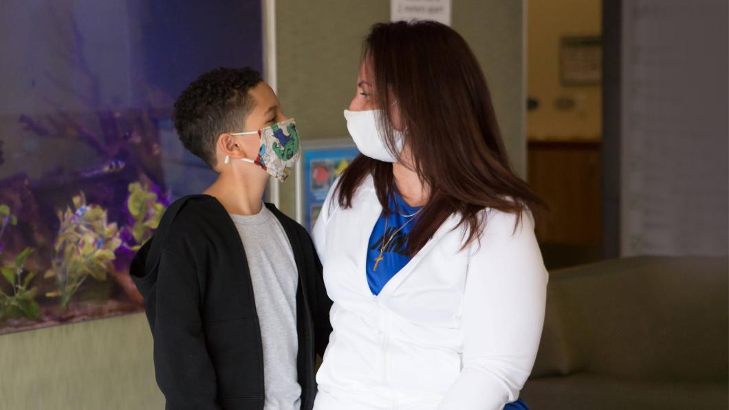 A child with medium-skin tone. They are wearing a mask, a black sweater and a grey t-shirt. The adult beside them has light skin-tone and brown hair. They are wearing a mask, a blue t-shirt and a white sweater.