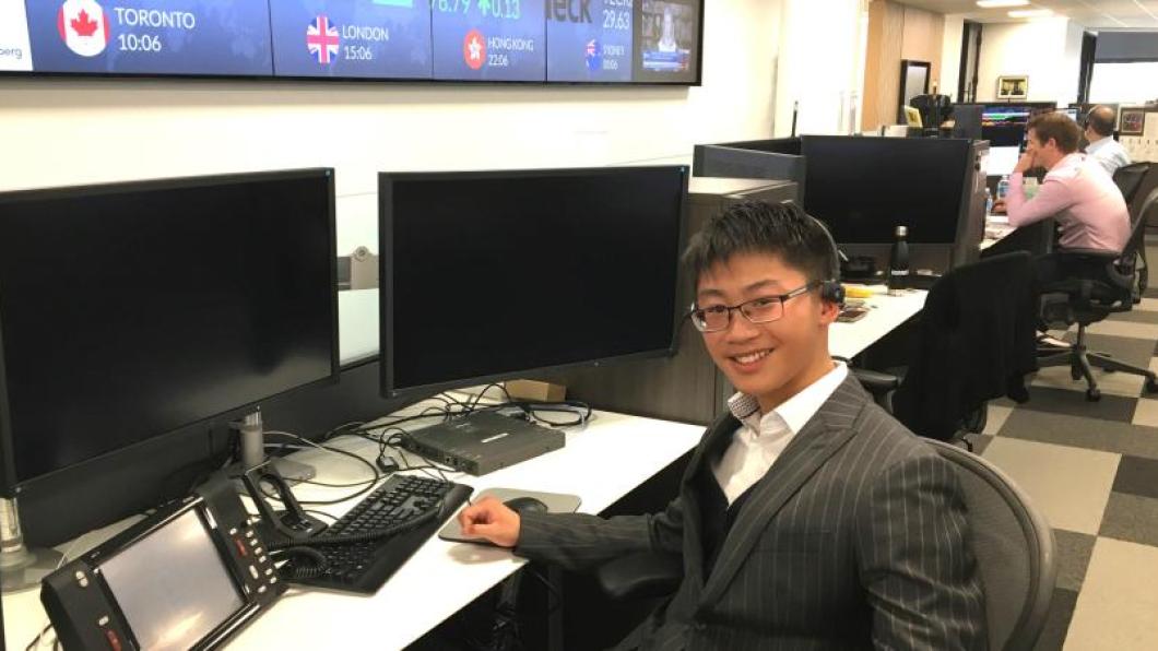 A young person sitting in front of a computer screen and smiling. They are wearing a black suit jacket with white stripes and black glasses.
