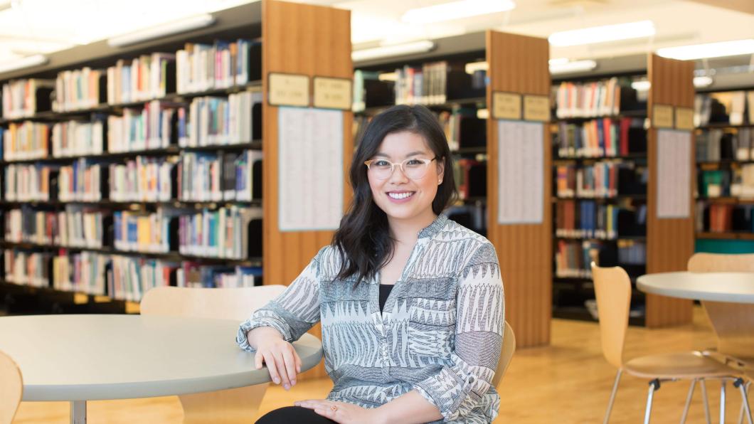 A woman with medium skin tone, long black hair and glasses. She is sitting in a library in front of several shelves of books.