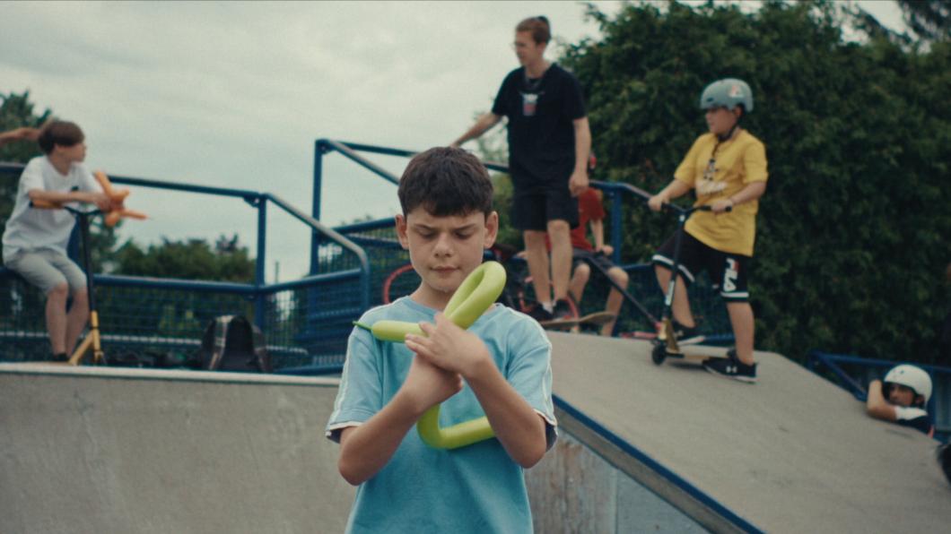 Boy making a balloon hat with boys standing on skateboard ramp behind