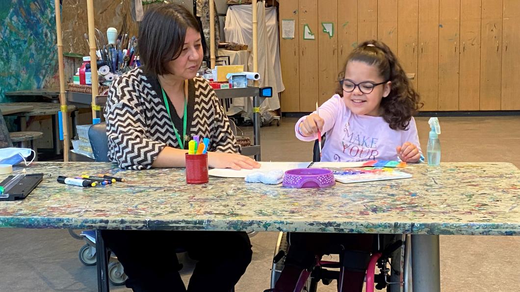 Child with dark hair in pink wheelchair sits at table painting with a woman beside her in an art studio