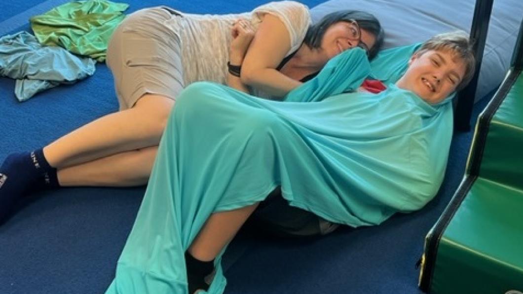 A woman and boy, wrapped in a turquoise blanket, lie on a blue mat