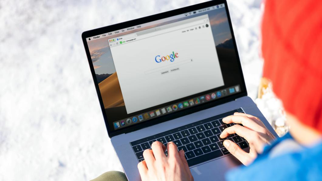 Person wearing a blue hoodie and orange toque is sitting with a grey laptop open and browsing on the Internet.