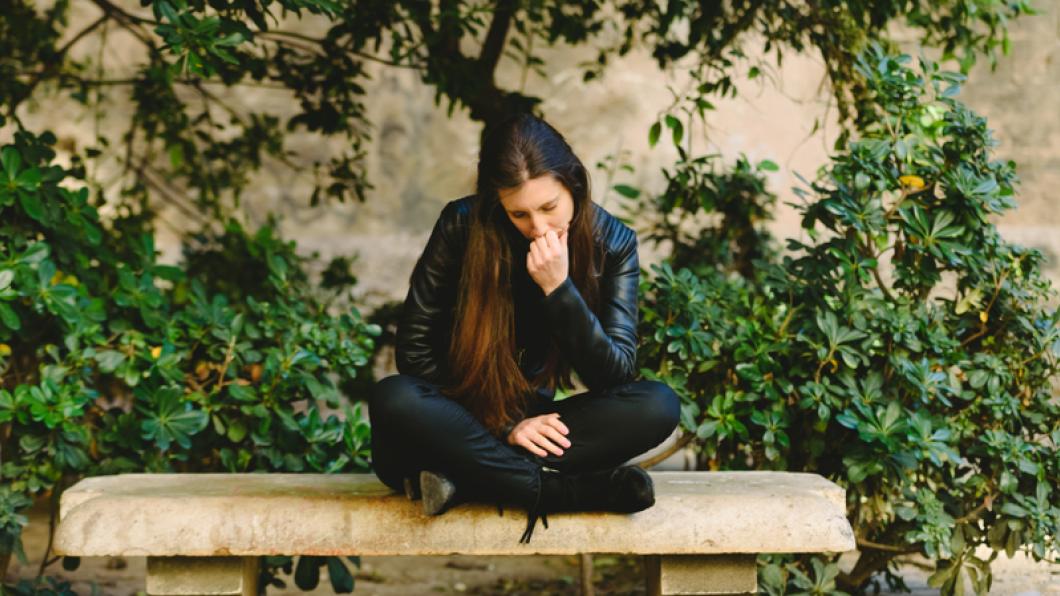 Young woman with long dark hair sits on bench with head down in her hand
