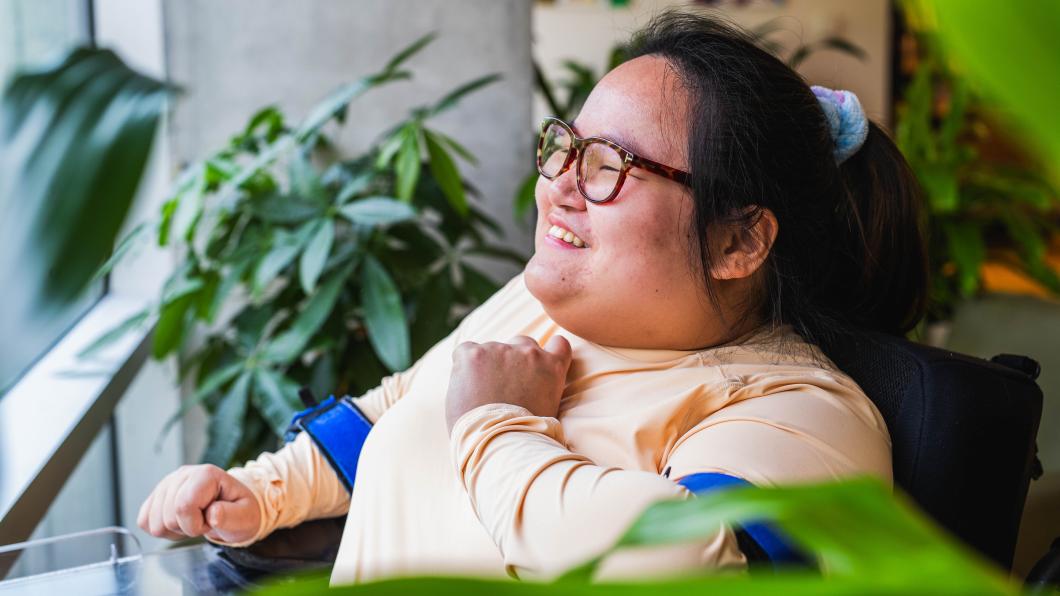A young woman who uses a powerchair smiles and looks out a window