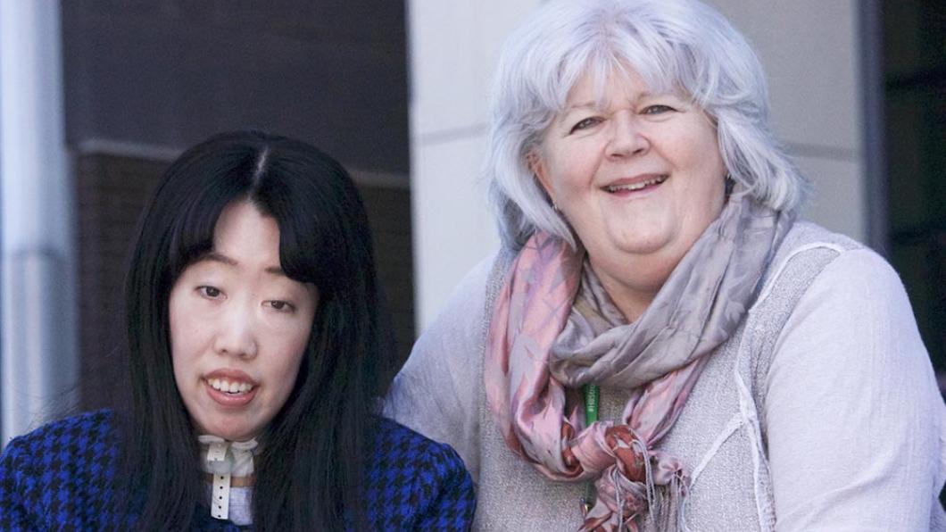 Woman with long black hair in navy sweater with a tracheotomy cork in her neck smiles with woman with grey hair wearing a pink and purple scarf