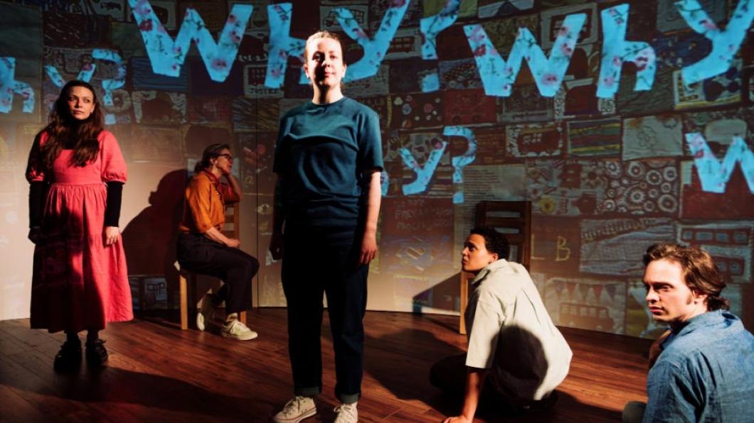 Actors standing and sitting on a stage with the word 'Why?' superimposed in blue on a screen behind them