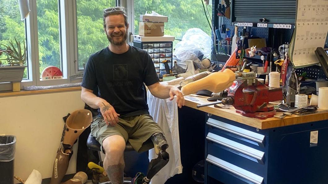 Man with beard and prosthetic leg sits on chair beside work table with tools