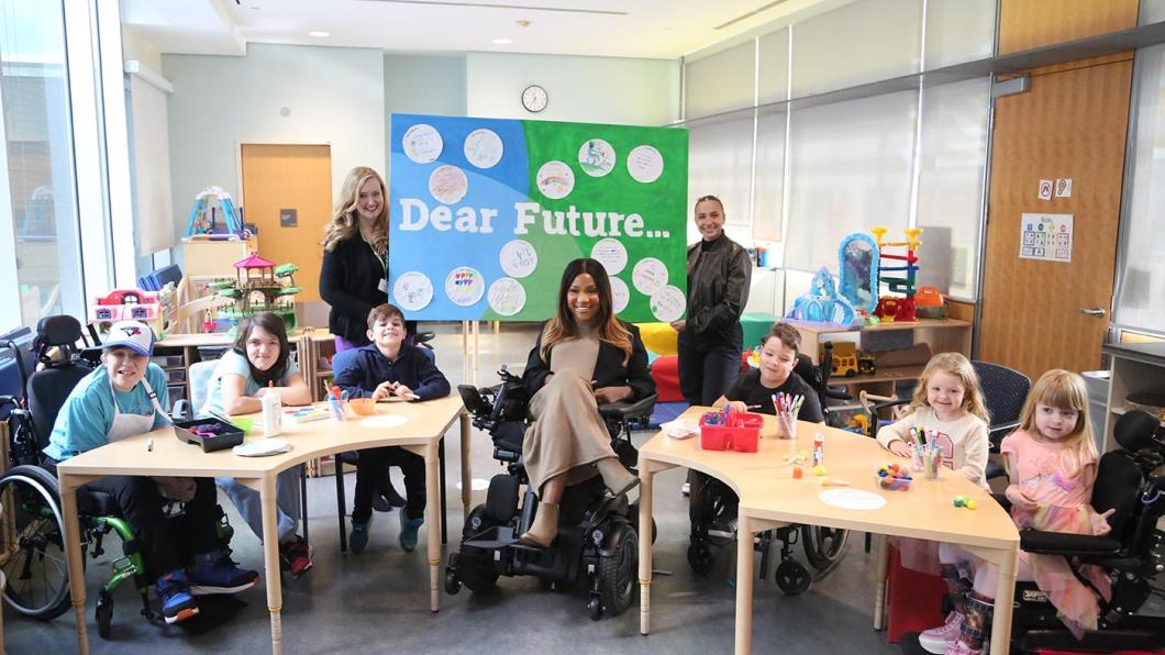 Children, teenagers and adults sitting and standing at a table of craft supplies. Two adults hold a green and blue banner that reads, 'Dear Future.'