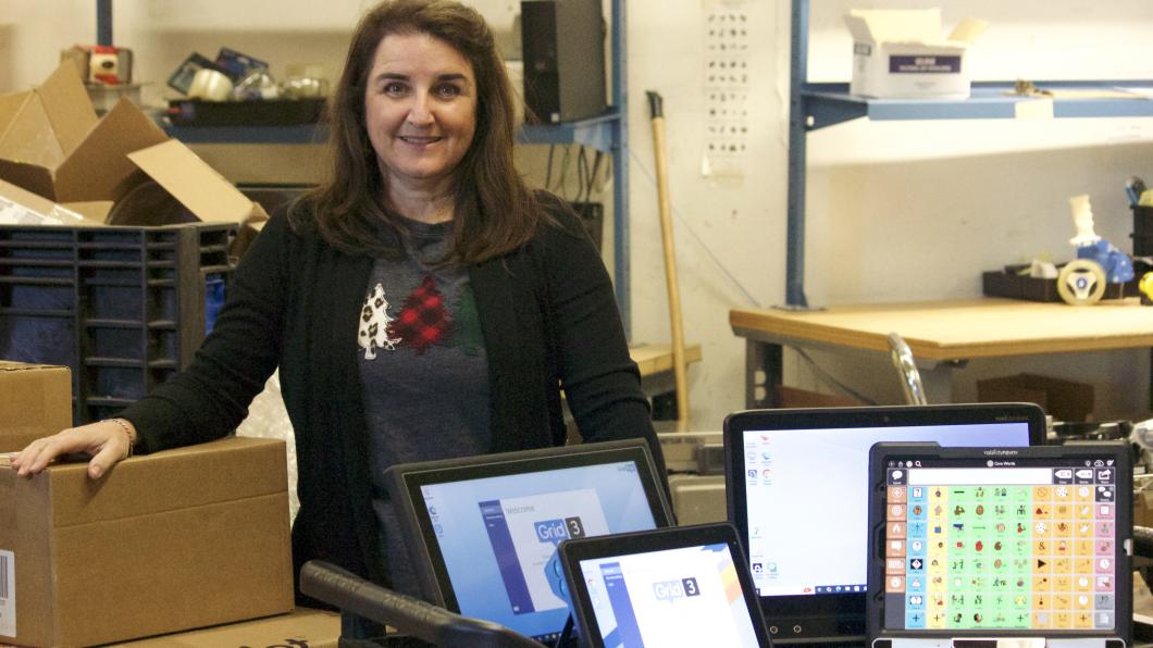 Woman with long hair stands in front of trolley with a number of computers displayed and boxes behind in warehouse