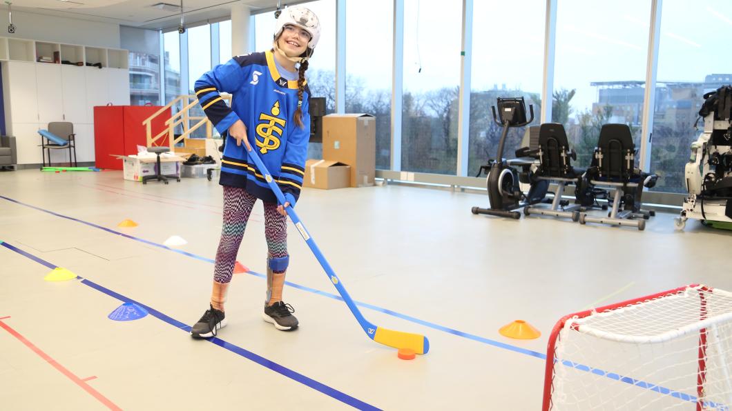 A child holding a hockey stick in front of a hockey net, wearing a helmet and ankle brace.