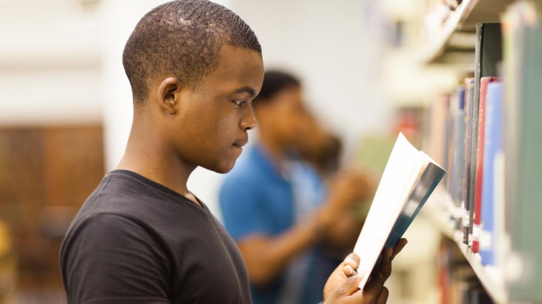 Young man in black t shirt with close cropped hair reads a publication in a library