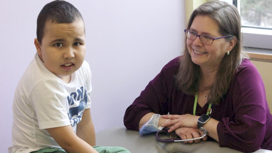 Boy with short dark hair and white t shirt sits on medical table with woman with long hair and glasses smiling at him