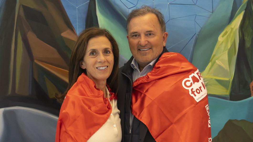 Two adults wearing red capes and standing in front of a colourful wall.