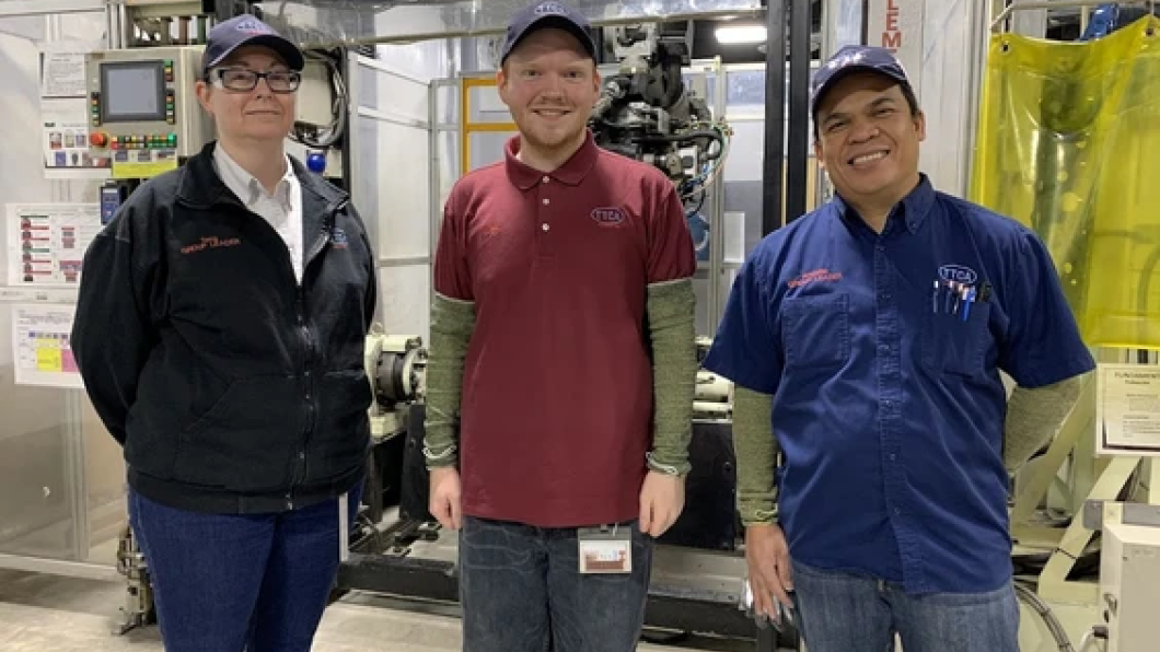 Alex stands in between two other adults in front of some factory equipment