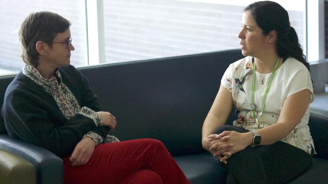 Woman with short hair and red pants sits on blue coach with another woman with white shirt and long dark hair