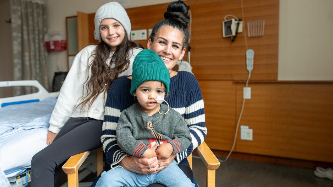 Two young children sit on their mom's lap, sitting on a chair in front of a hospital bed