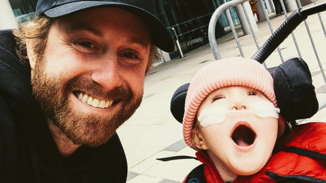 Man in baseball cap smiles beside young girl in stroller wearing nasal prongs