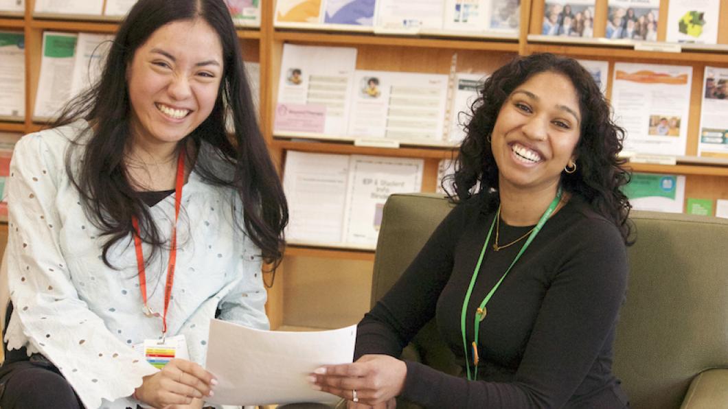Two women with dark hair hold papers with shelves behind them