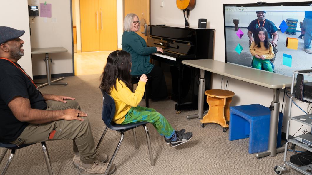Child in yellow shirt sits in front of screen with icons with a woman on piano beside her and a man sitting behind her