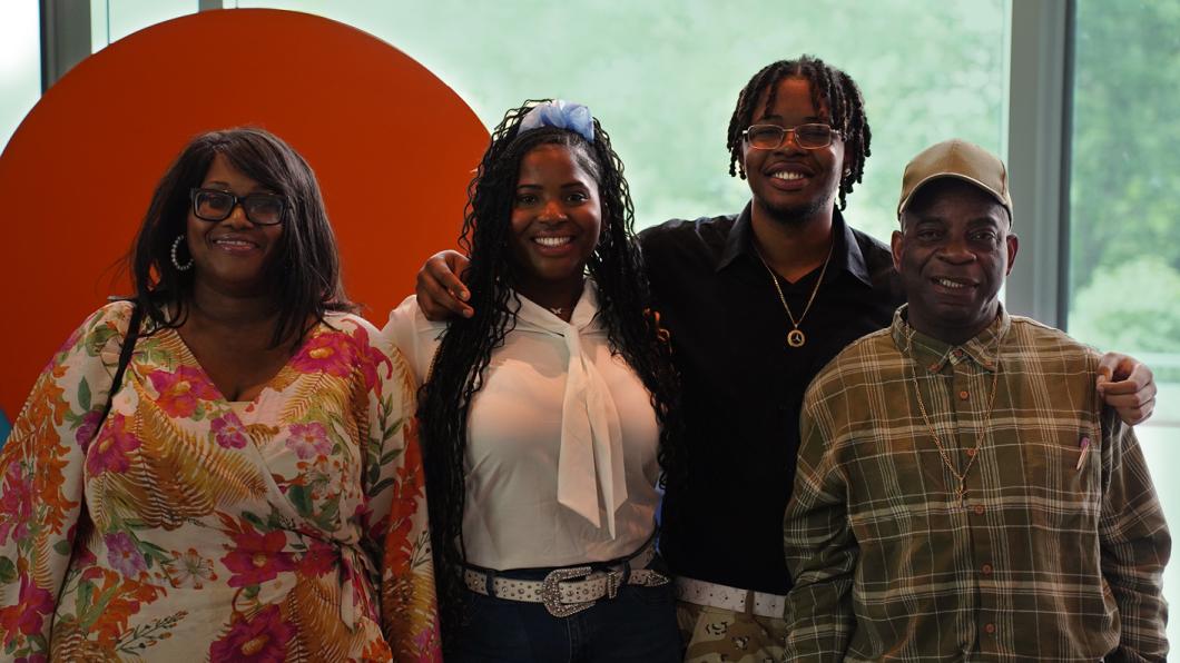 Photograph of four people standing side by side in front of a large orange circular backdrop with a green blurred background. They are wearing casual clothing, 
