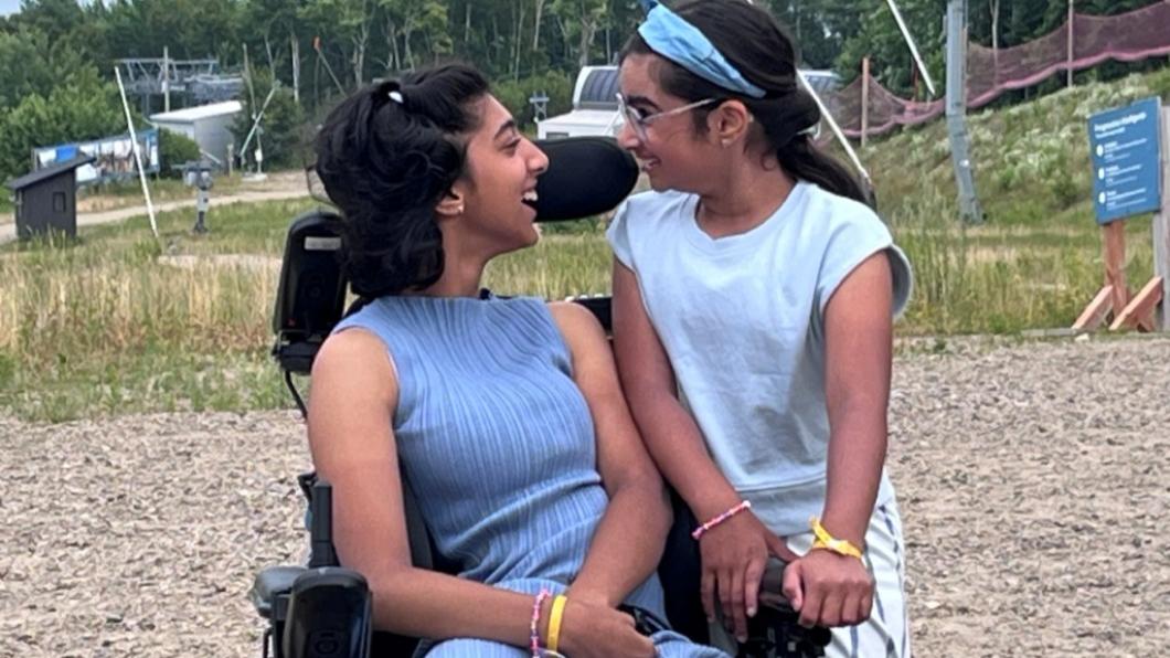 Teen girl in wheelchair and lavender outfit smiles at girl with bandana standing with arms on her chair