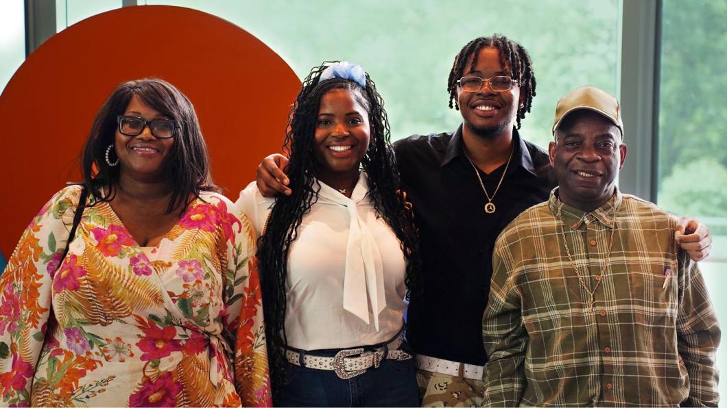 Photograph of four people standing side by side in front of a large orange circular backdrop with a green blurred background. They are wearing casual clothing, 