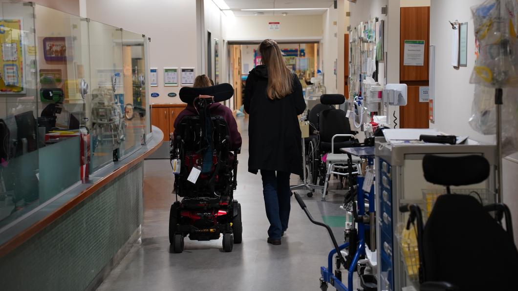 Young woman in wheelchair and woman with blonde hair walk through a hospital corridor