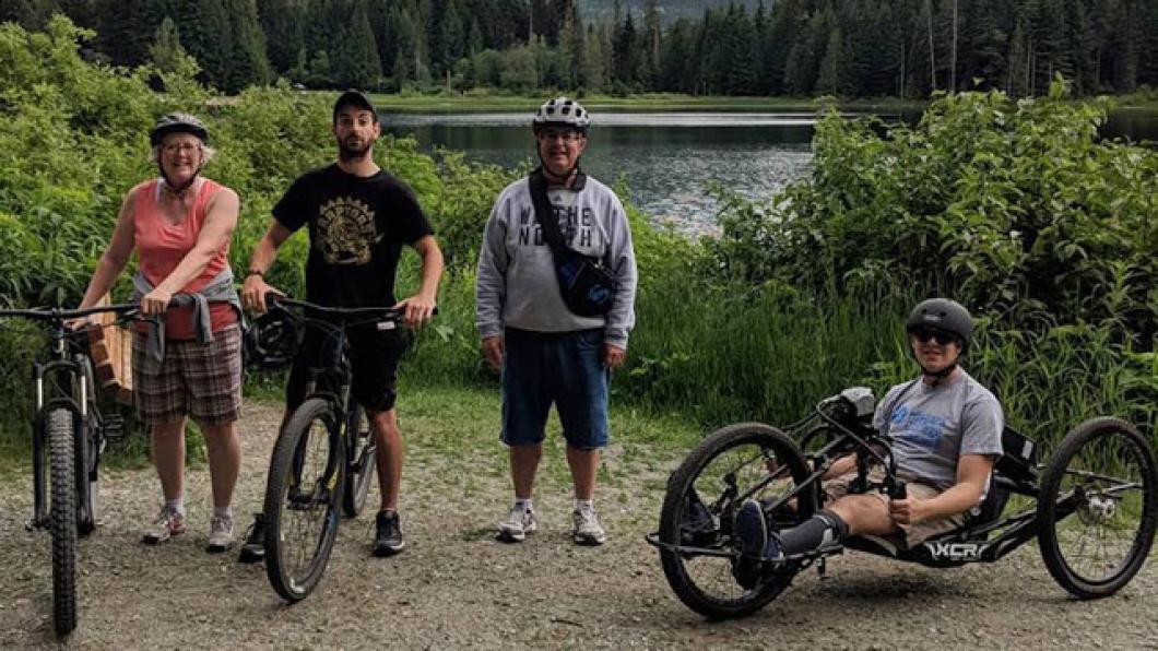 A woman standing next to a bile, man standing next to bike, man in adaptive bike and a man standing 