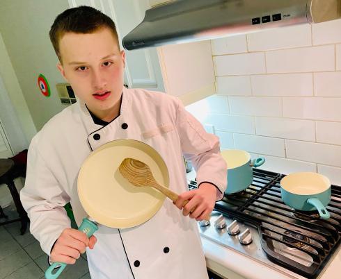 Mick in a chef's jacket holding up a pan and spoon in a kitchen.