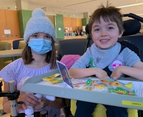 A young boy with light skin tone and short brown hair. He is using a wheelchair and a tray, which has school supplies and activities on it. A child with light skin tone, medium-length brown hair is kneeling beside him. They are wearing a knit hat and a blue face mask. 