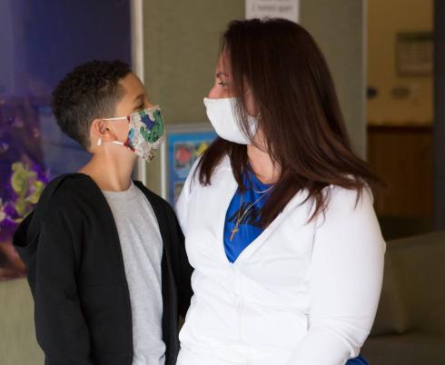 A child with medium-skin tone. They are wearing a mask, a black sweater and a grey t-shirt. The adult beside them has light skin-tone and brown hair. They are wearing a mask, a blue t-shirt and a white sweater.