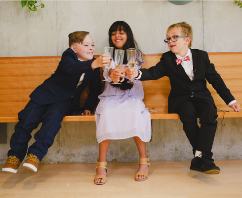 Three children sitting on a brown indoor bench. The first child has light skin tone, brown hair and is wearing a blue suit. The second child has medium skin tone, long black hair and is wearing a white dress. The third child has light skin tone, short blonde hair and is wearing glasses and a black suit. The children are toasting their drinks.