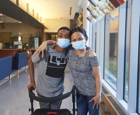 Image of a young adult wearing a grey t-shirt and standing behind a mobility device with their arm around another adult