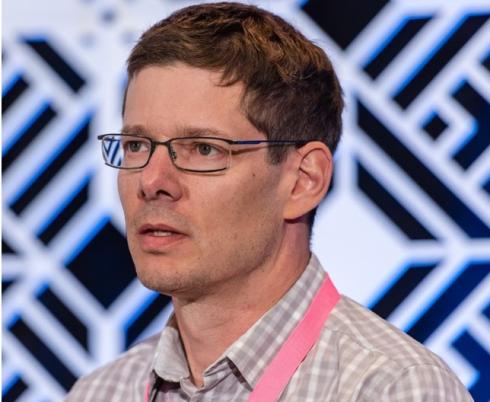 Dr. Jaimie Borisoff stands against a black and white checkered back drop wearing a light pink button up shirt, black glasses and short brown hair