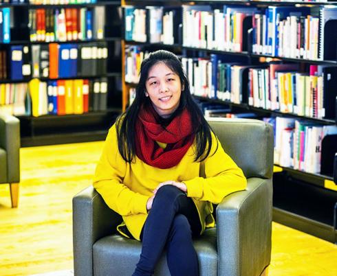 Youth leader Lexin sits in a chair surrounded by shelves of books. She is wearing a yellow sweater and a red scarf.