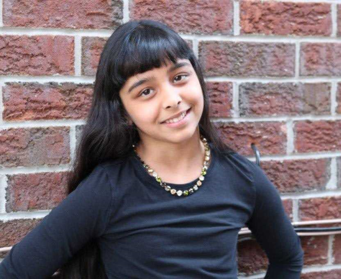 A young girl with long black hair and medium skin tone. She is smiling while standing in front of a red-brick wall, posing with her hands on her hips.