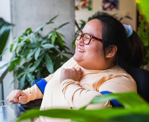 A young woman who uses a powerchair smiles and looks out a window